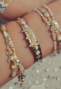 Trois bracelets sur un poignet : un bracelet jonc en or avec des motifs d'oiseaux et des perles, une chaîne tressée en or et blanc, et un bracelet en perles avec des pendentifs.