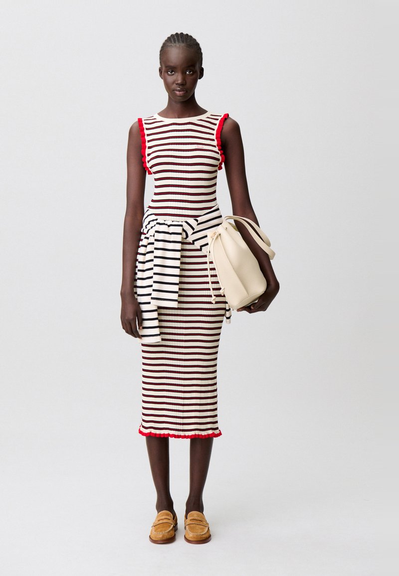 Abito aderente a righe con dettagli a volant rossi, abbinato a un maglione a righe bianco e nero annodato in vita. Borsa beige e mocassini.