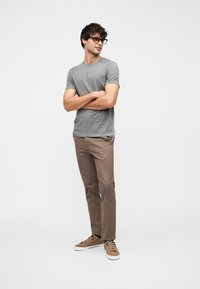 Jeune homme portant des lunettes, bras croisés, vêtu d'un t-shirt gris, d'un pantalon marron et de baskets beiges sur un fond blanc uni.
