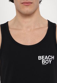 Jeune personne portant un débardeur noir avec le texte "BEACH BOY" imprimé en blanc sur la poitrine, montrant le cou et le haut du torse.