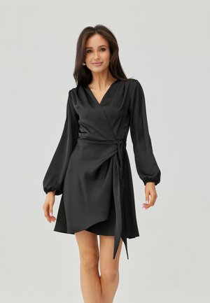 Žena v černých dlouhých rukávech, zavinovacích šatech se zavazováním na boku, stojící před jednoduchým světle šedým pozadím.