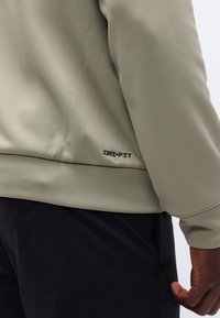 Κοντινό πλάνο ατόμου που φορά μπεζ μπουφάν Dri-FIT και μαύρο παντελόνι, δείχνοντας το κάτω μέρος της πλάτης και το δεξί χέρι με ορατό κείμενο της μάρκας.