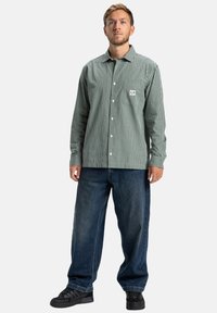 Ein Mann steht in einem grün karierten Hemd mit Knöpfen, lockeren blauen Jeans und schwarzen Turnschuhen vor einem schlichten weißen Hintergrund.