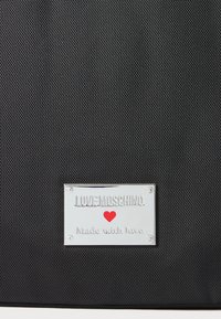 Čierna textúrovaná tkanina so striebornou tabuľkou s nápisom "LOVE MOSCHINO" a červeným srdcom nad textom "Vyrobené s láskou".