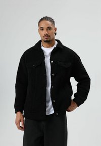 Zwarte sherpa-jas met knoopsluiting, twee voorzakken, gestructureerde stof en een kraag, gedragen over een wit T-shirt.