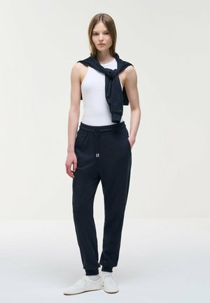 Giovane donna che indossa una canottiera bianca, pantaloni della tuta neri, scarpe da ginnastica bianche e una felpa nera legata intorno alle spalle, in piedi.