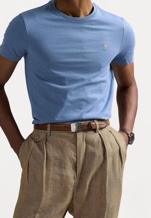 Mężczyzna ubrany w jasnobeżową bluzkę z krótkim rękawem i beżowe, plisowane spodnie w kratę z brązowym paskiem, zegarkiem i rękami w kieszeniach.