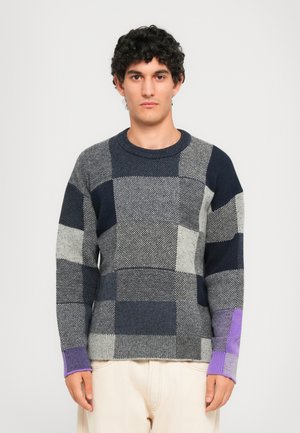 Man in een marineblauw, grijs en paars geblokt vest met crèmekleurige broek, staande tegen een effen lichtgrijze achtergrond.