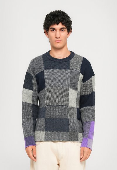 Ein Mann trägt einen navy-, grau- und lila-karierten Pullover mit cremefarbenen Hosen und steht vor einem schlichten hellgrauen Hintergrund.