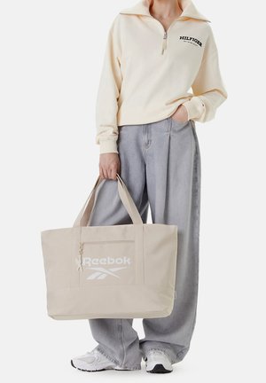 Persona che indossa una felpa crema con cerniera e jeans grigi a gamba larga, che tiene in mano una grande borsa a tracolla beige Reebok con logo bianco.