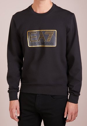 Osoba w czarnej koszulce z długim rękawem z złotym logo EA7 Emporio Armani na piersi.
