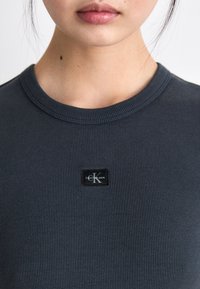 Tamsiai pilkas raukšlėtas marškinėliai su apvalia iškirpte ir maža juoda etikete su užrašu "Calvin Klein Jeans" bei baltu "K" logotipu.