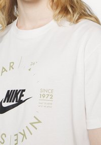 T-shirt en coton blanc avec un logo Nike noir et du texte vert, comprenant "DEPUIS 1972" et des coordonnées géographiques. Design classique à col rond.