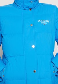 Blå puffjacka med hög krage, med en glänsande yta, tryckknappar och en vit "tommy jeans"-logotyp på framsidan.