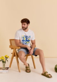 Witte graphic t-shirt met blauw en groen ontwerp, denim shorts en olijfgroene sandalen, zittend op een houten stoel met gele bloemen.