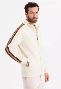 Veste zippée blanche avec des accents rayés marron sur les manches, les poches latérales et les poignets côtelés. Tissu doux avec un design à col haut.