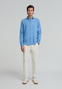 Cămașă albastră deschis, din denim, cu guler standard și mâneci lungi, asociată cu pantaloni crem și adidași albi.