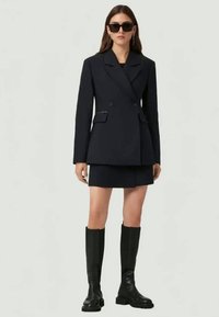 Blazer bleu marine à double boutonnage avec revers, mini-jupe assortie et bottes noires à hauteur du genou. Le mannequin est accessoirisé avec des lunettes de soleil surdimensionnées.