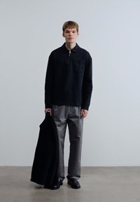 Μαύρο πόλο με μακριά μανίκια, φερμουάρ στο λαιμό και τσέπη, συνδυασμένο με γκρι παντελόνια φαρδιάς γραμμής και μαύρα παπούτσια, κρατώντας ένα σκούρο παλτό.