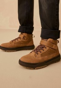 Braune Wildlederstiefel mit einem hohen Design, ausgestattet mit dunkelbraunen Schnürsenkeln und einem Klettverschluss, auf einer strukturierten schwarzen Gummisohle.