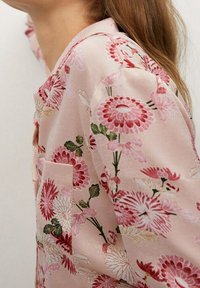 Chemise à motif floral en tissu rose pâle, avec une poche, abondance de fleurs rouges et blanches, et des feuilles vertes. Texture lisse, coupe décontractée.