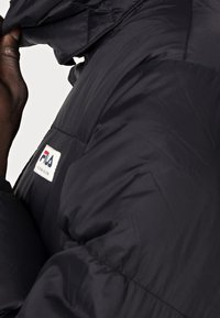 Giacca a vento nera con colletto alto, dotata di una piccola etichetta bianca con logo. Il tessuto ha una texture liscia e un dettaglio di cuciture notevole.