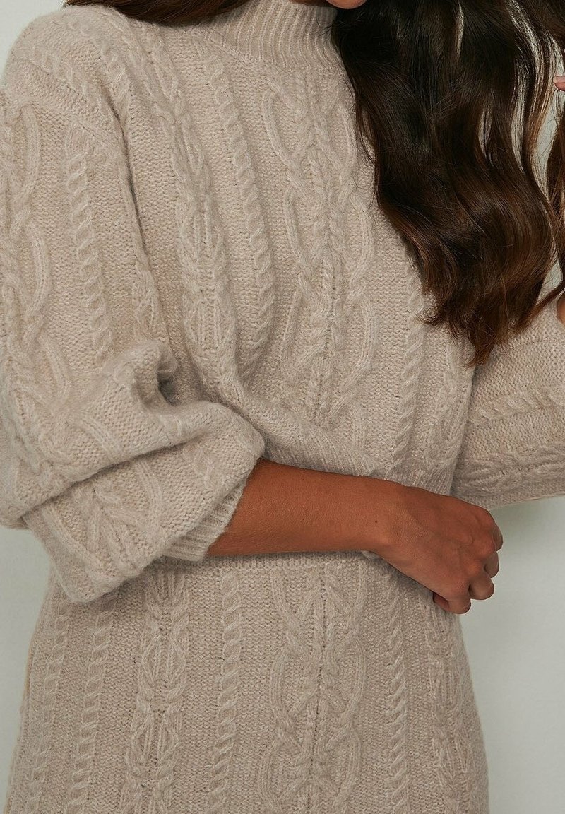 Pull beige en maille torsadée avec col montant, motif texturé et manches larges retroussées. Le tissu en maille douce accentue l'apparence chaleureuse.