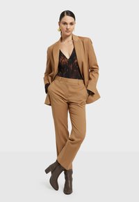 Smeđi odijelo u prugama s krojenim sakoom i odgovarajućim hlačama. Ispod se nalazi tamna majica s suptilnim apstraktnim uzorkom. Smeđe čizme do članka.