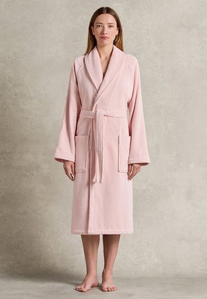 Donna in piedi scalza che indossa un accappatoio rosa chiaro fino al ginocchio, con maniche lunghe, colletto a scialle, cintura e due tasche frontali.