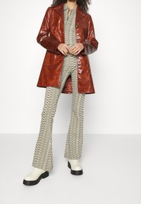 Brun glansig läderjacka med spetsig krage och knappar, lager-på-lager över beige mönstrade utsvängda byxor, kombinerat med vita platåstövlar.