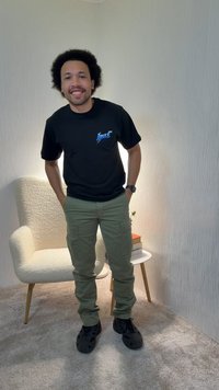 Mann mit Afro-Frisur, der ein schwarzes T-Shirt mit Grafik und grüne Cargohosen trägt, steht in einem mit Teppich ausgelegten Raum neben einem weißen Sessel und einem kleinen runden Tisch.