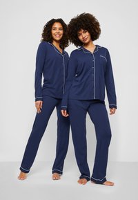 Marinblå pyjamasset i mjukt tyg med en knapp-up lång ärmad topp med vit piping och matchande byxor med långa ben och uppvikta fållar.