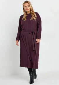 Lång, räfflad klänning i djup lila med rund halsringning och bälte i midjan. Långa ärmar och en avslappnad passform. Bärs med åtsittande svarta stövlar.