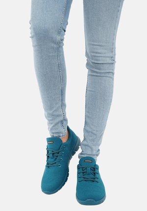 Turkusowe sneakersy na sznurówki na osobie noszącej jasnoniebieskie dopasowane dżinsy, pokazujące buty i dolne części nóg na białym tle.
