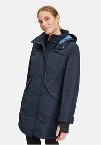 Marineblau wasserdichte Jacke, bis zur Mitte des Oberschenkels, mit Kapuze und Druckknopfverschluss. Verfügt über Reißverschlusstaschen und kontrastierende blaue Akzente.
