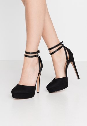 Black platform heels with a suede material, featuring patent ankle straps, a slender high heel, and a rounded toe design.