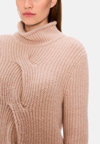 Rosa gestrickter Pullover mit hohem Kragen, der ein strukturiertes Muster und eine asymmetrische Aussparung an der Vorderseite aufweist. Weiches, flauschiges Material.