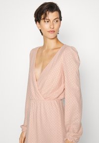 Vestido cor pêssego com um padrão texturizado, decote em V profundo e mangas longas com punhos franzidos, apresentando uma cintura ajustada e saia fluida.