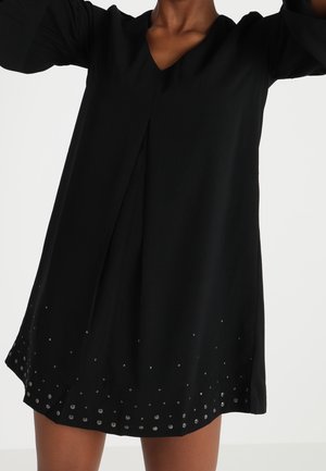 Femme portant une robe noire à manches longues avec un col en V et des clous argentés décorant l'ourlet, les bras levés au-dessus de la tête, fond blanc uni.