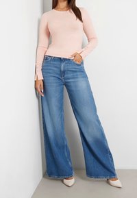 Vaaleanpunainen pitkähihainen paita, jossa on nappikoristeet kalvosimissa, yhdistettynä leveälahkeisiin sinisiin farkkuihin ja kermaan vivahtaviin korkokenkiin.