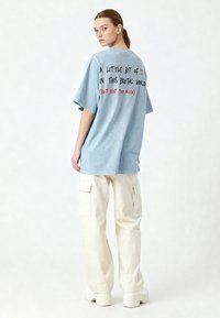 Světlomodré oversized tričko s textem na zádech: "MALÝ KOUSEK ❤️ V TOMTO BRUTÁLNÍM SVĚTĚ (ALE NE PŘÍLIŠ)". Text je v černé a červené.