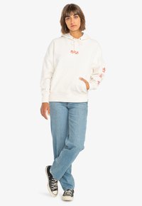 Cream hoodie with a front pocket and drawstring, featuring red graphic prints on sleeves. Paired with light blue jeans and black sneakers.