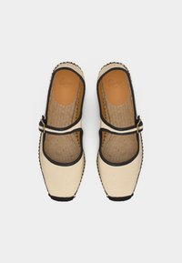 Paire de chaussures Mary Jane beiges avec bordures et boucle noires, semelles intérieures texturées et bouts carrés sur fond blanc.