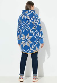 Felpa con cappuccio blu a motivi floreali e accenti beige, dotata di maniche lunghe e orlo leggermente curvato. Abbinata a jeans scuri.