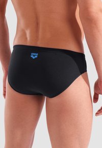 Uomo che indossa un costume da bagno nero di Arena, visto di spalle, su uno sfondo grigio chiaro neutro.