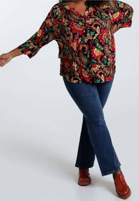 Blusa floreale colorata con sfondo nero, scollatura a V, maniche a 3/4 e orlo arricciato, abbinata a jeans di denim a zampa e scarpe rosse.