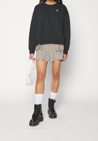 Felpa nera con logo bianco, pantaloni corti beige a pieghe con tasche, stivali neri con lacci e calzini bianchi alla caviglia, che tiene una piccola borsa bianca.