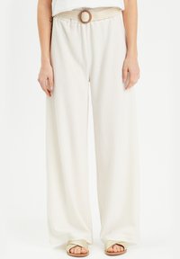 Pantalones anchos blancos y texturizados con un cinturón trenzado que presenta una hebilla circular. Combinados con sandalias planas de colores claros.
