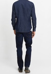 Tmavá džínová košile s dlouhými rukávy, zapínáním na knoflíky na manžetách a zadním dílem. Kombinováno s tmavými slim fit džínami a černými botami.