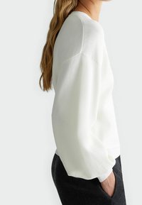 Femme portant un haut texturé à manches longues blanches avec des coutures d'épaule tombantes et un pantalon noir, debout avec une main dans la poche.